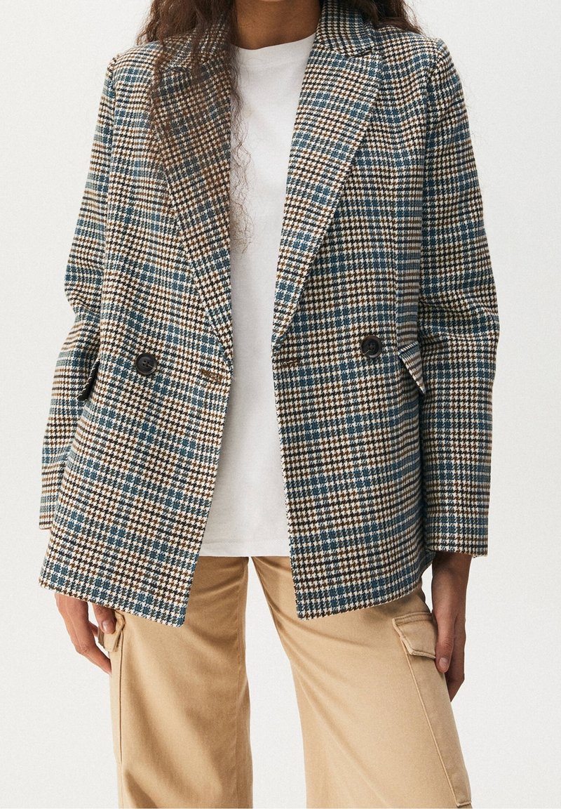 Femme portant un blazer pied-de-poule marron, bleu et blanc sur une chemise blanche avec un pantalon cargo beige, debout sur un fond uni.