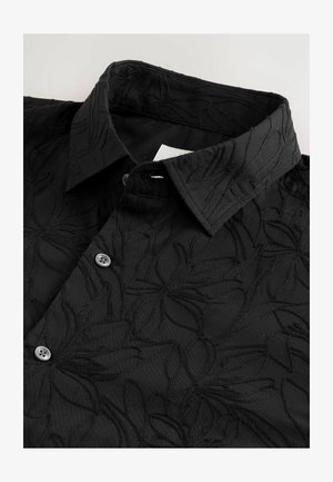 Chemise noire avec un motif floral brodé, dotée d'un col pointu et d'une fermeture par bouton. Le tissu a une finition texturée et mate.