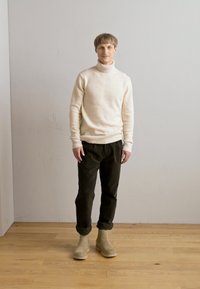 Suéter de cuello alto crema, pantalones verde oscuro y botas de tobillo color beige claro; de pie sobre un suelo de madera contra un fondo sencillo.
