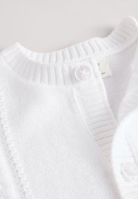 Cardigan blanc en tricot avec col côtelé et boutons transparents sur fond clair.