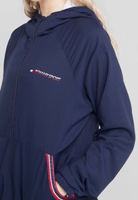 Marinblå lättviktsjacka med huva, med dragkedja i fronten, elastiska ärmslut och Tommy Sport-logotyp i rött och vitt på bröstet och fickorna.