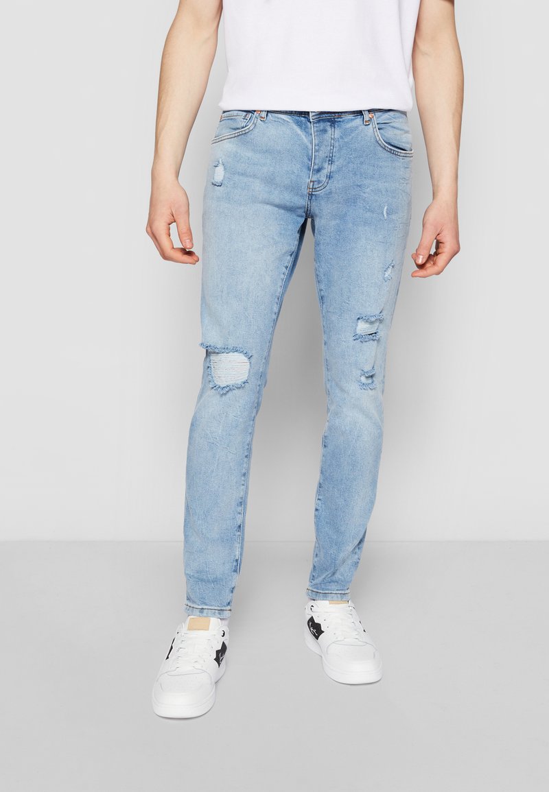 Svetlo modri skinny jeans z raztrganimi detajli, ki vključujejo strgane punkcije in zbledeli videz. V kombinaciji z belo majico in supergami.