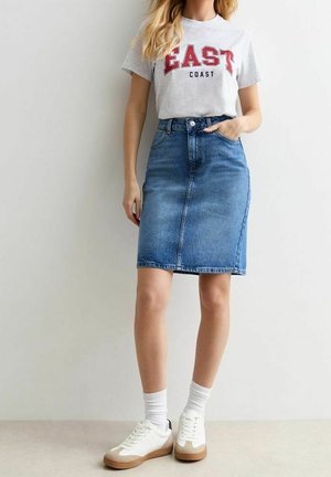 Femme portant un t-shirt blanc avec un texte rouge "EAST COAST", une jupe en jean bleue longueur genoux, des chaussettes blanches et des baskets blanches.