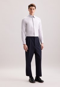 Jonge man staat, gekleed in een wit overhemd met knopen, marineblauwe geplooide broek, zwarte sokken en zwarte nette schoenen op een effen achtergrond.