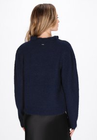 Dunkelblaues Strickpullover mit einem hohen Kragen, lockerer Passform und leicht strukturiertem Stoff. Verfügt über ein kleines Logo-Detail am hinteren Kragen.