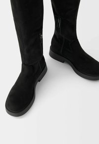 Svarta mockakängor i knähöjd med en slät textur, utrustade med sidodragkedjor och en platt gummisula. Enkel design utan mönster.