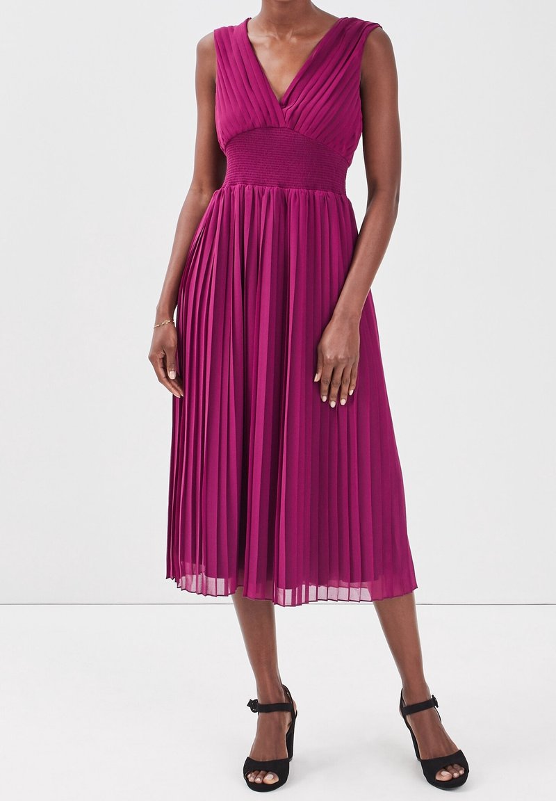 Vestido fucsia con pliegues, escote en V profundo, cuerpo texturizado y falda fluida, combinado con tacones negros de tiras al tobillo.