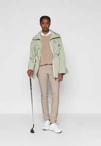 Ανοιχτό πράσινο μπουφάν με φερμουάρ, με σφηκοφωλιά στη μέση και δύο τσέπες μπροστά, σε συνδυασμό με μπεζ πουλόβερ και στενά μπεζ παντελόνια. Άσπρα αθλητικά παπούτσια και μπαστούνι γκολφ.