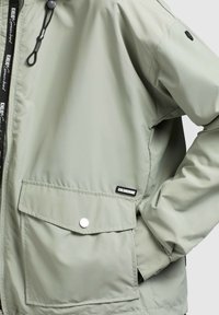 Veste gris clair avec poche à rabat devant, bouton-pression, fermeture éclair et étiquettes de marque noires et blanches sur la poitrine et près de la poche.