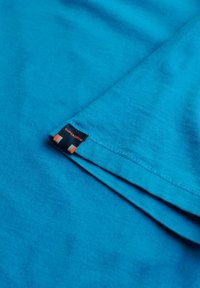 Turkis bomulds T-shirt med korte ærmer. Har et blåt og orange mærke i kanten. Glat tekstur med en ensartet overflade.