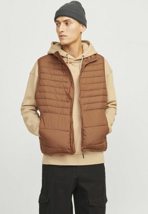 Brun puffer vest med vandret syninger, lynlås foran, båret over en tan hættetrøje. Mørk hue. Sorte cargo bukser.