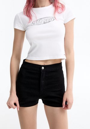 Kvinde med pink hår iført en hvid cropped t-shirt med teksten "not you" og højtaljede sorte shorts, stående foran en hvid baggrund.