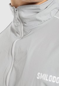 Primo piano del colletto di una giacca zip-up grigia chiara con zip visibile e parte del testo ricamato in bianco "SMILODO".