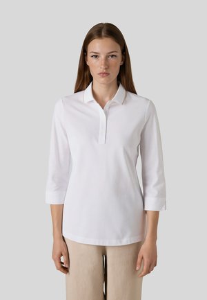 Femme aux cheveux bruns et raides portant un polo blanc à manches longues et un pantalon beige, debout devant un fond gris uni.