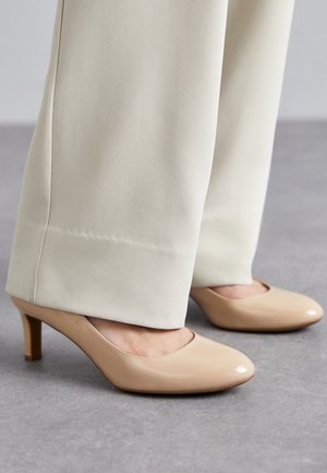 Chaussures basses en cuir verni beige portées avec un pantalon crème à jambes larges sur un sol gris.
