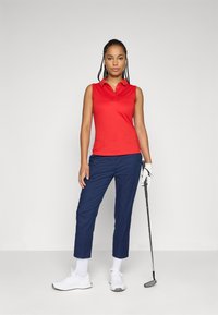 Donna in polo rosso senza maniche e pantaloni blu navy, che tiene una mazza da golf con un guanto bianco, in piedi contro uno sfondo chiaro e uniforme.