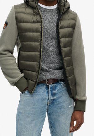 Mężczyzna w oliwkowo zielonej pikowanej kurtce z rękawami z polaru, szarym dzianinowym swetrze, niebieskich dżinsach i brązowym pasku, z rękami częściowo w kieszeniach.