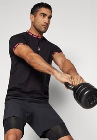 Black athletic t-shirt with red logo and trim, paired with black shorts featuring textured accents. Individual holds a kettlebell.