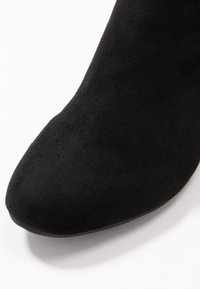 Bota de tobillo de ante negro con una textura suave, punta afilada y un tacón delgado. El enfoque está en la parte superior del zapato.