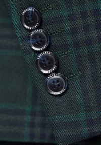 Zielono-niebieska tkanina w kratę z czterema czarnymi guzikami wyraźnie wyeksponowanymi, z tekstem "LANCERTO" wokół dziurek na guziki.