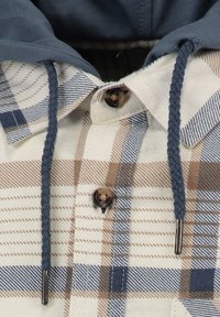 Camicia a quadri con linee beige, marroni e blu. Materiale in misto cotone con cappuccio e lacci intrecciati. Bottoni marroni decorano la parte anteriore.