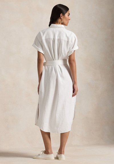 Femeie în picioare, purtând o rochie midi albă cu curea, mâneci suflecate și tăieturi laterale, asortată cu adidași albi, fundal bej.