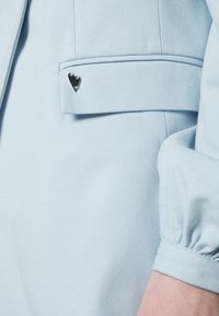 Blusa azul clara com uma textura suave, apresentando um pequeno detalhe em forma de coração em prata no bolso e punhos franzidos.