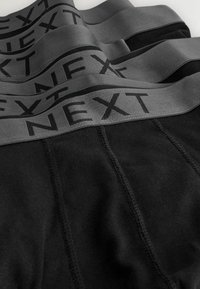 Tela negra con anchas bandas grises en la cintura que presentan la palabra "NEXT" en letras negras y en negrita, en varias prendas dobladas.