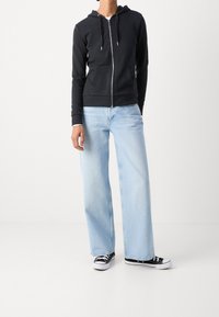 Schwarze Zip-Hoodie mit Kapuze und Fronttaschen, kombiniert mit hellblauen weit geschnittenen Jeans und schwarzen Stoffschuhen mit weißen Sohlen.