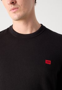 Pulover negru cu guler rotund, fabricat dintr-un material moale, având un mic logo dreptunghiular roșu pe pieptul stâng. Design simplu, clasic.