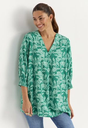 Smilende kvinde med mørkt hår samlet i en hestehale, iført en grøn blomstret V-hals bluse med trekvartlange ærmer og blå jeans, stående op ad en hvid væg.