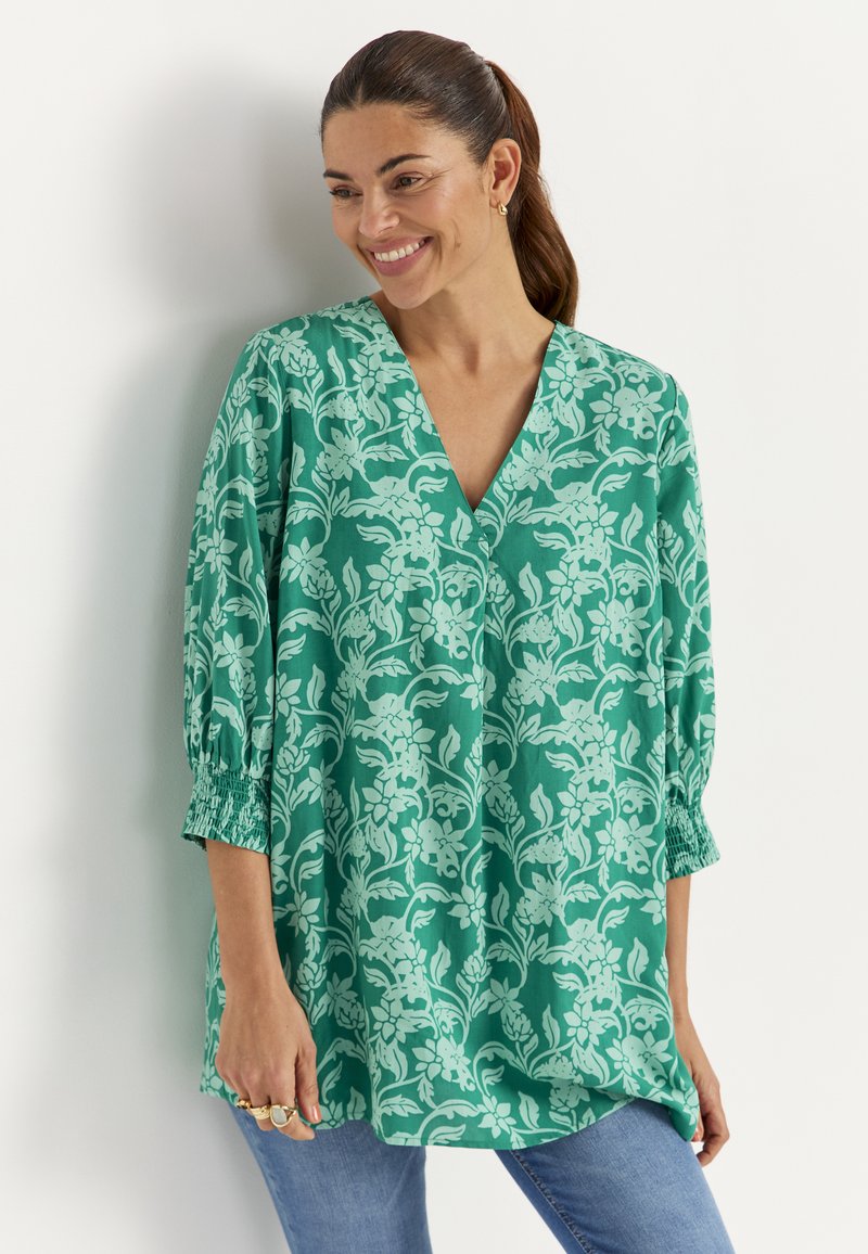 Glunderende vrouw met donker haar in een paardenstaart draagt een groene blouse met bloemenprint, V-hals en driekwart mouwen, gecombineerd met een blauwe spijkerbroek, staand tegen een witte muur.