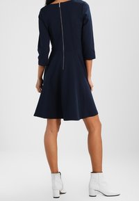 Granatowa sukienka z rozkloszowanym dołem, rękawami o długości trzech czwartych i zamkiem błyskawicznym z tyłu pośrodku. Noszona z białymi botkami za kostkę.