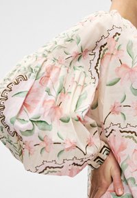Manche bouffante avec motif floral rose et feuilles vertes sur tissu blanc, portée par une personne avec la main posée sur la hanche.