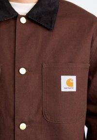 Brown canvas jacket with a black corduroy collar, featuring a front pocket and a square logo patch in orange and white. Gold button detail.