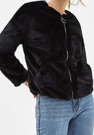 Woman wearing a black fuzzy zip-up jacket with pockets and light blue jeans, standing with one hand on her hip.