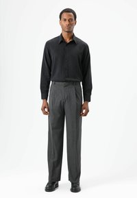 Zwarte blouse met lange mouwen en knopen, gecombineerd met grijze, geplooide broek. De outfit heeft een gladde textuur en een klassieke pasvorm, aangevuld met zwarte schoenen.