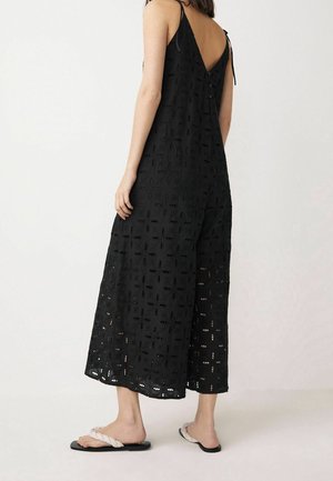 Black eyelet fabric jumpsuit with wide legs, V-back design, and adjustable spaghetti straps. Model wears flat braided sandals.