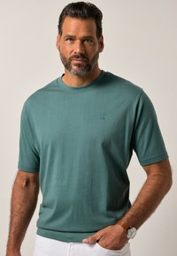 T-shirt a maniche corte di colore teal in morbido cotone, con scollo a girocollo e logo discreto. Il modello indossa pantaloni bianchi e un orologio marrone.