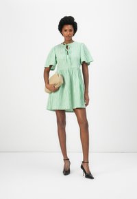 Vestido riscado em verde e branco com mangas curtas e franzidas, decote com abertura e saia evasê, combinado com uma mala bege e sapatos de salto pretos.