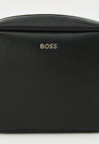 Juodas odinis kosmetikos krepšys su tekstūruotu pavirščiu. Priekyje yra auksinis "BOSS" logotipas ir šiek tiek apvalus viršus.
