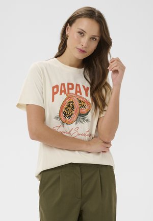 Mladá žena s dlhými hnedými vlasmi, oblečená v bielom tričku s grafickým motívom papáje a olivovo zelených nohaviciach, pózujúca pred jednoduchým pozadím.