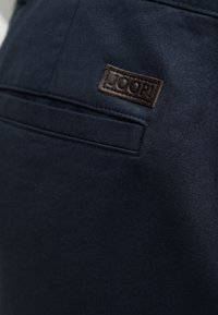 Κοντινή λήψη σκούρου ναυτικού υφάσματος που δείχνει μια οριζόντια τσέπη με τρύπα και ένα μικρό ορθογώνιο δερμάτινο έμπλαστρο με την επωνυμία «JOOP!».