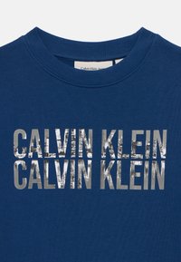 Blå hettegenser med rund hals, med en iøynefallende sølvfarge print av "CALVIN KLEIN" på forsiden, laget av et mykt, teksturert stoff.
