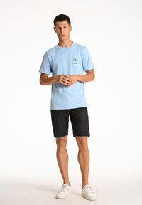 Vaaleansininen lyhythihainen t-paita, jossa on pieni palmun brodeeraus, yhdistettynä mustiin shortseihin ja valkoisiin lenkkareihin, tasaisen taustan edessä.