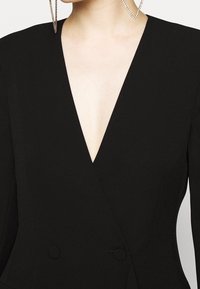 Robe portefeuille noire avec un décolleté en V profond, dotée de doubles boutons, d'épaules structurées et d'une texture de tissu lisse.