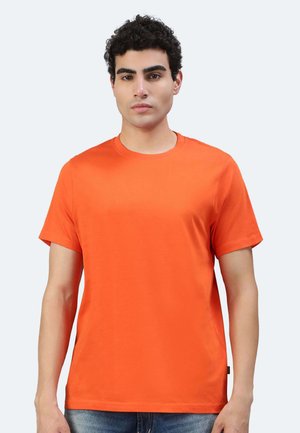 Uomo con capelli ricci che indossa una t-shirt arancione a maniche corte semplice e jeans blu, in piedi davanti a uno sfondo grigio chiaro.