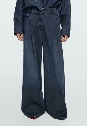 Personne portant un pantalon large en denim foncé et une chemise à manches longues assortie en denim foncé, les mains relâchées le long du corps sur un fond uni.