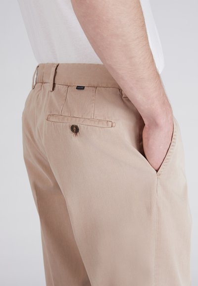 Pantaloni in cotone beige con una tasca posteriore con chiusura a bottone e un'etichetta sulla vita, mostrati da un'angolazione laterale.