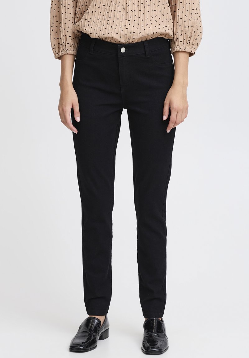 Femme portant un pantalon noir moulant, un chemisier beige à manches longues avec de petits motifs noirs, et des mocassins noirs vernis, debout sur fond blanc.